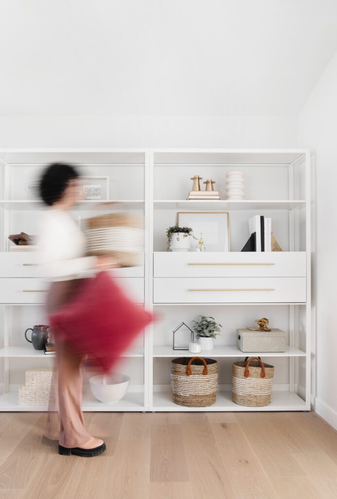 A blurred person walking through a living room which is styled with shelves and baskets. The person is carrying a red cushion