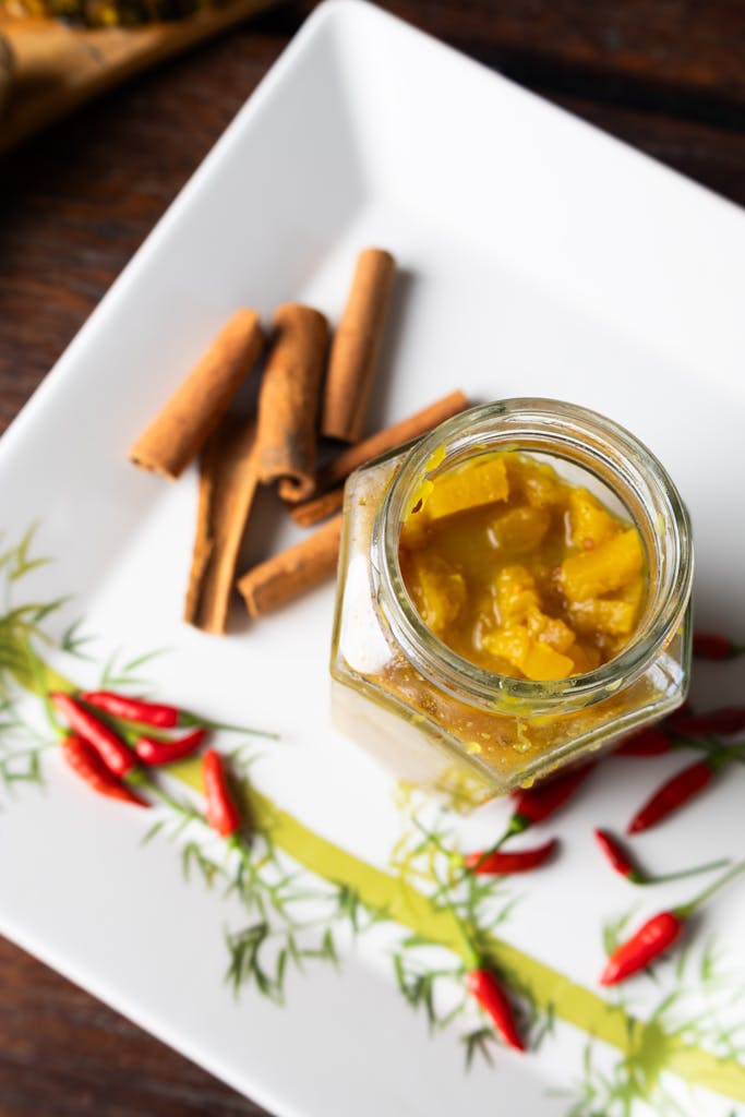 Top view of homemade mango chutney in a glass jar with cinnamon sticks and red chillies on a decorative plate.