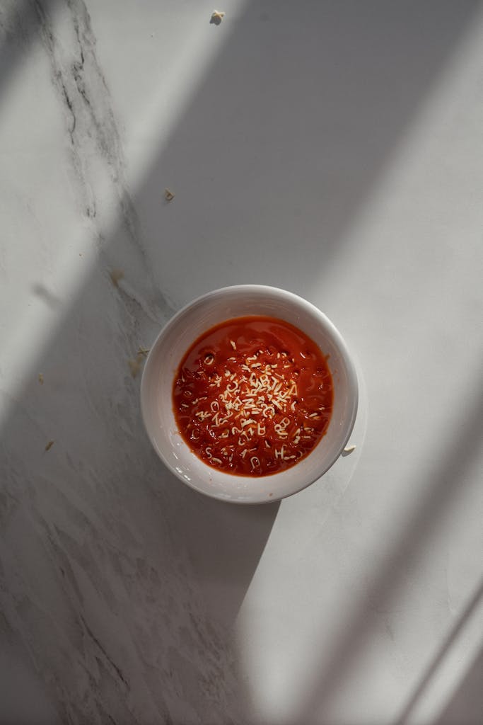 A ceramic bowl filled with tomato soup topped with shredded cheese, casting shadows on a marble surface.