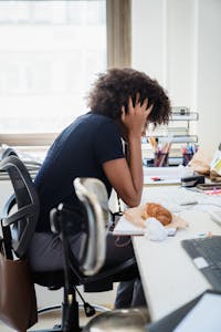 A woman in an office holds her head in stress, croissant on desk.