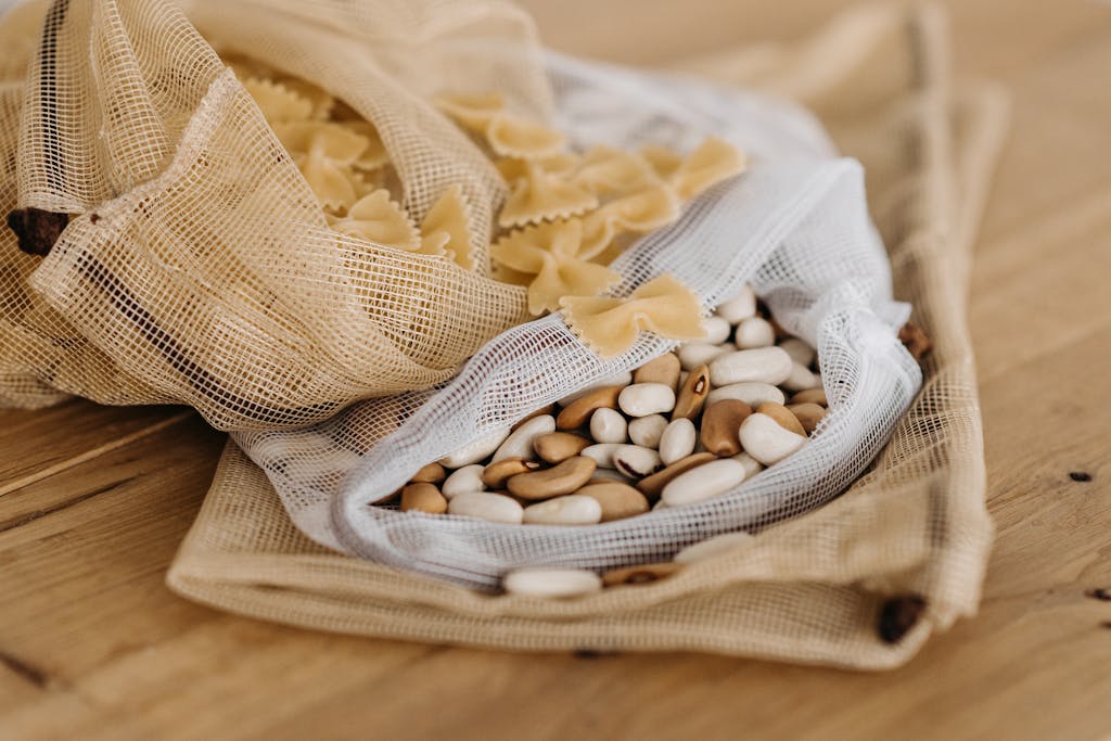 Eco-friendly bags filled with raw farfalle pasta and mixed beans on a wooden surface.