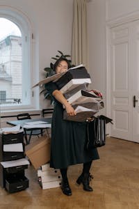 Asian woman in a black dress struggling to hold multiple document boxes in an office setting.