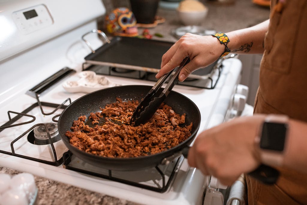 Hands cooking chilli meat in a pan on a gas stove with tongs.